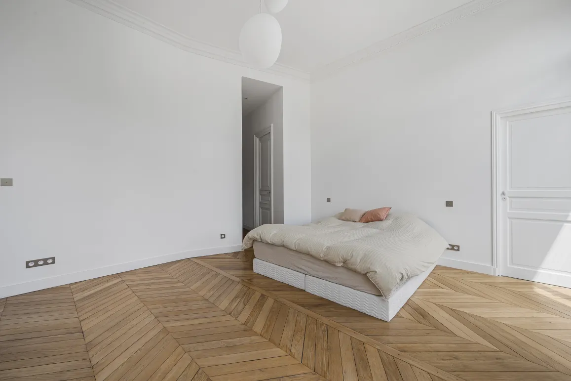 Chambre luxe minimaliste, parquet chevron, murs blancs. Élégance parisienne, luminosité naturelle. Idéal appartement haussmannien.