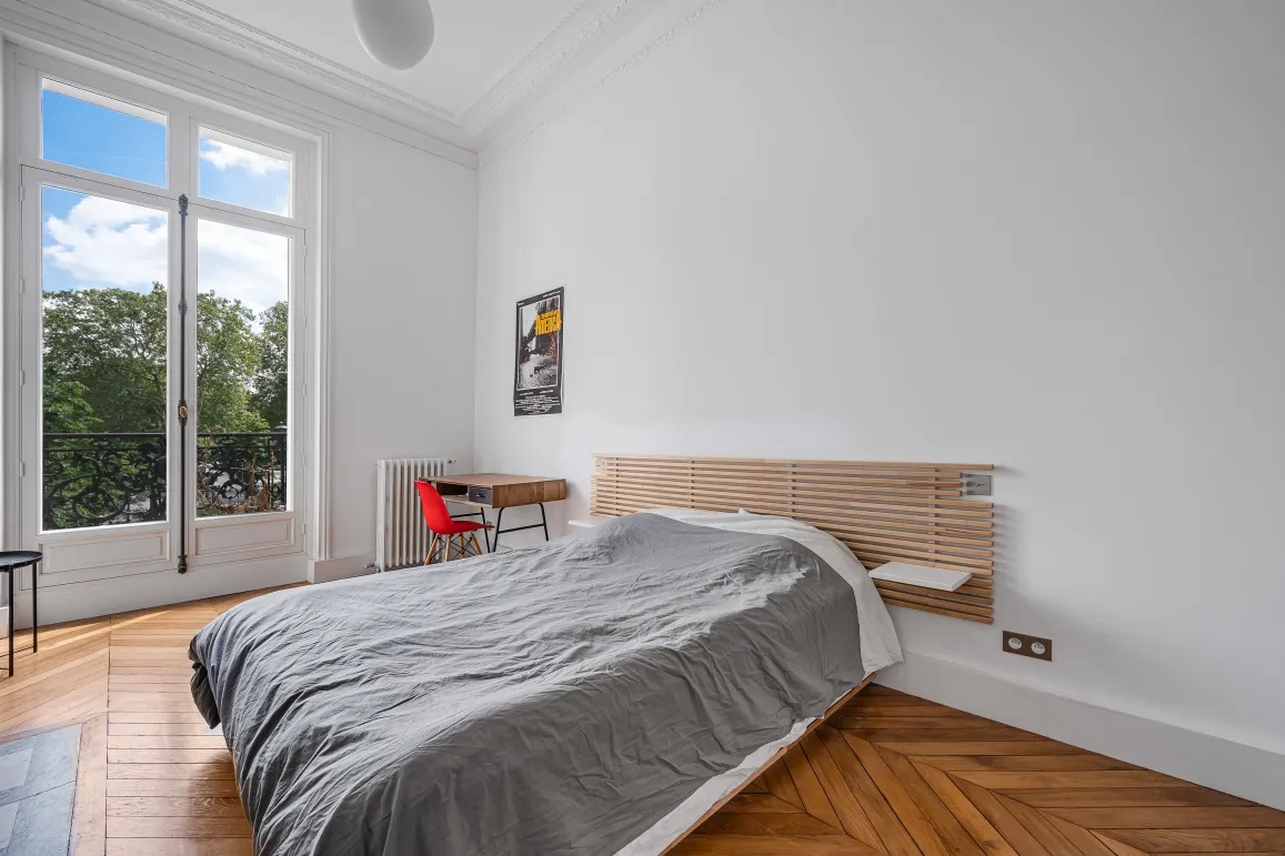 Chambre lumineuse avec balcon vue verdure, parquet chevrons et moulures. Design moderne et élégance parisienne pour un luxe discret.
