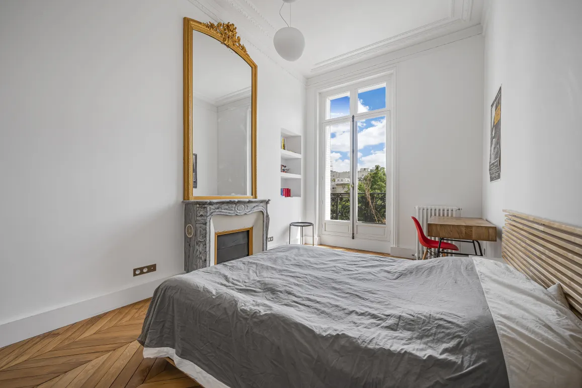 Chambre lumineuse avec parquet chevrons, cheminée marbre, miroir doré et vue dégagée. Charme haussmannien pour appartement de luxe.