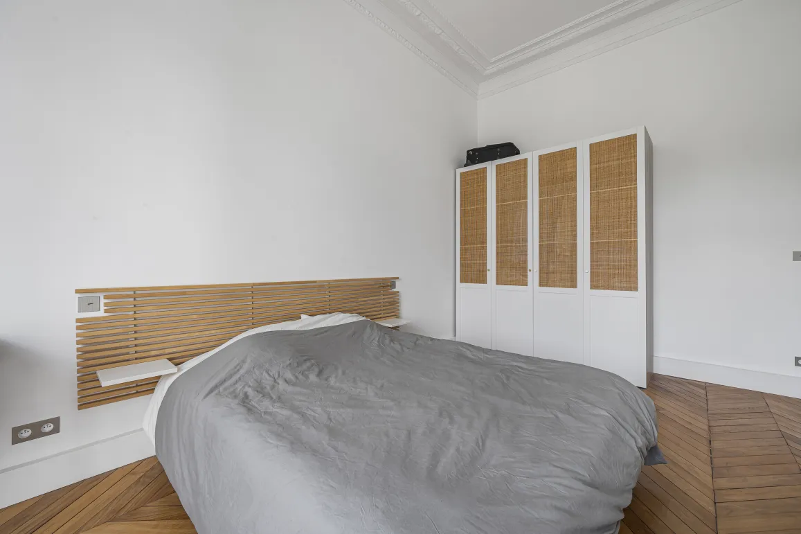 Chambre épurée: tête de lit bois, dressing rotin, parquet chevrons. Ambiance sereine et élégante pour un intérieur de luxe parisien.