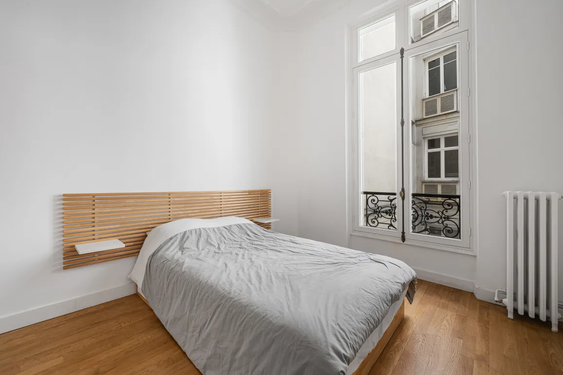 Chambre lumineuse au design épuré, parquet, tête de lit bois moderne, fenêtre parisienne. Appartement de prestige.