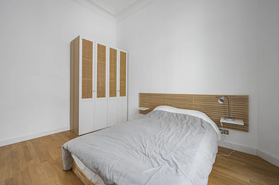 Chambre moderne et lumineuse avec parquet, tête de lit design en bois, et armoire élégante en rotin. Idéal pour appartement de luxe.