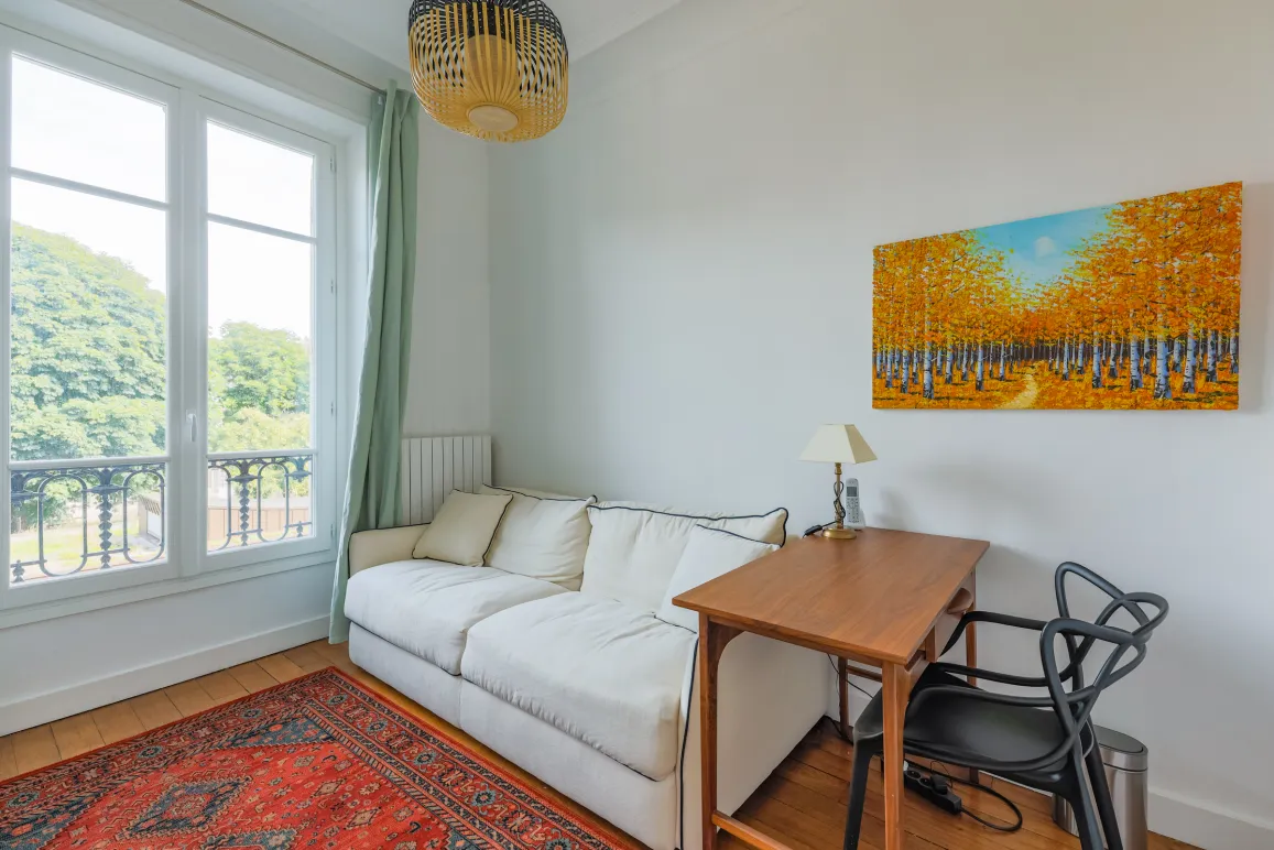 Pièce lumineuse avec vue verdoyante, canapé blanc, bureau bois et art moderne. Ambiance sereine et élégante pour travail ou détente.