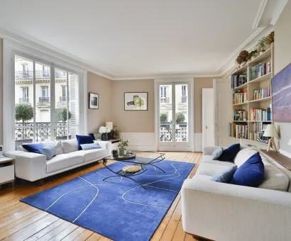 Salon de luxe parisien lumineux. Canapés blancs, tapis bleu moderne, parquet. Vue sur la ville. Élégance et confort haussmannien.