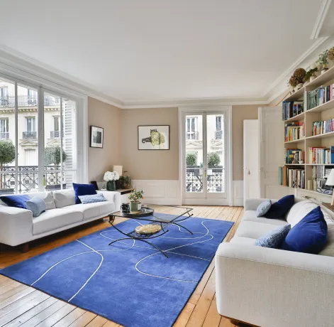 Salon de luxe parisien lumineux. Canapés blancs, tapis bleu moderne, parquet. Vue sur la ville. Élégance et confort haussmannien.