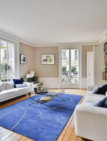 Salon de luxe parisien lumineux. Canapés blancs, tapis bleu moderne, parquet. Vue sur la ville. Élégance et confort haussmannien.